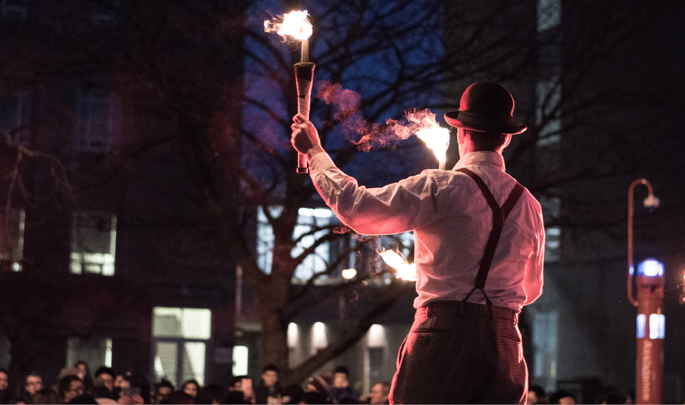 A nighttime image from behind of a person on stilts breathes fire on campus.