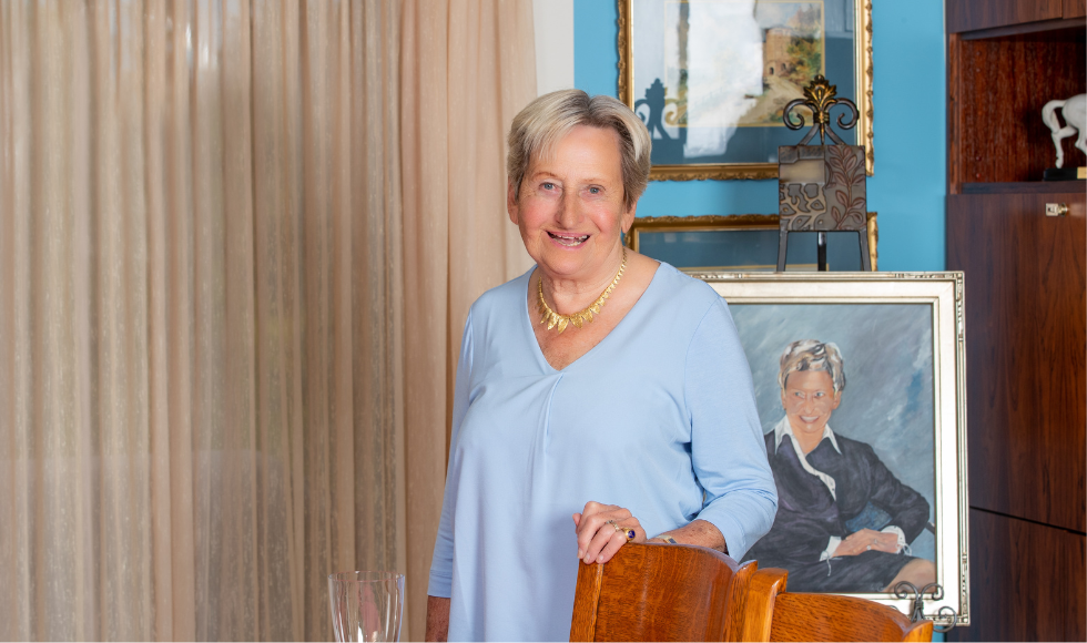 Margaret Juravinski has her hand on the top of a chair back and is standing in front of a painted portrait of herself.