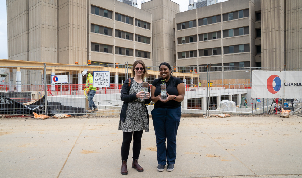 Two people standing in front of a construction site holding glass cases with plants growing in them