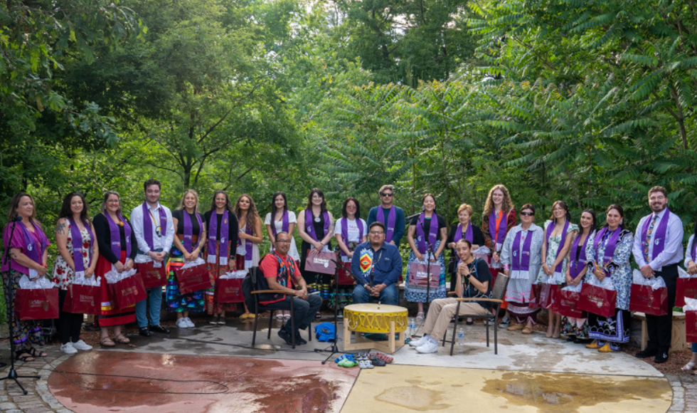 A group photo of graduating students wearing their purple stoles in a semicircle around the traditional drummers at the outdoor classroom.