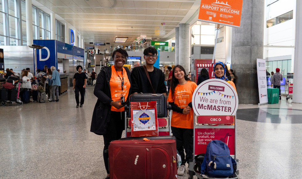 An arriving student with his cart full of luggage smiles with volunteers at the airport, beside a big 
