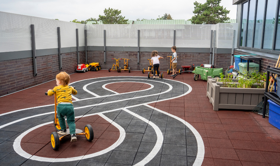 Three young children play on scooters in an outdoor terrace area