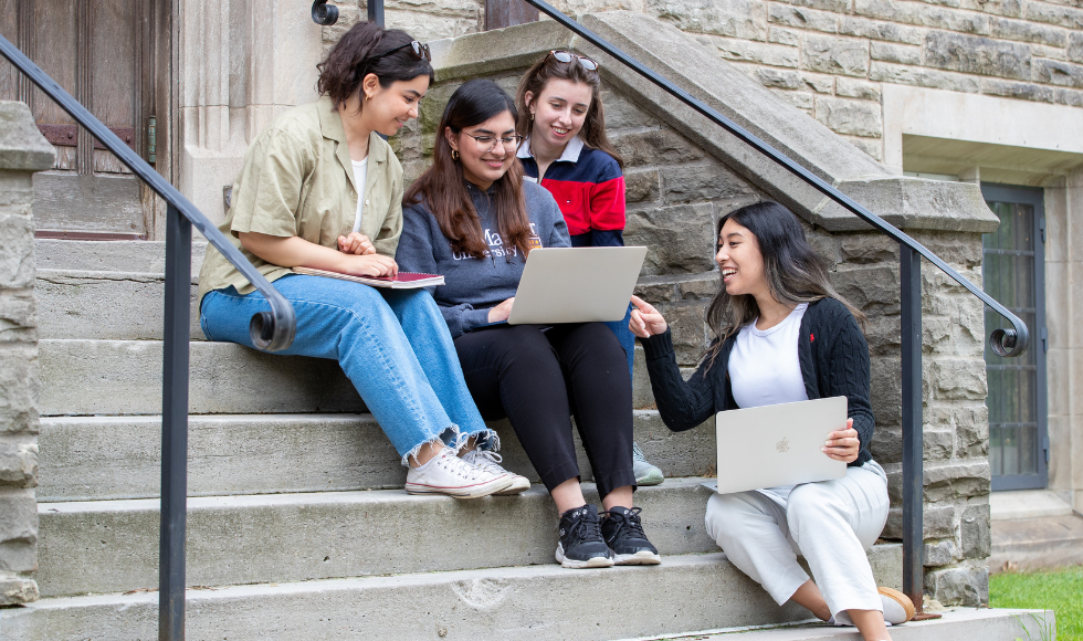 Four students, two with open laptops, sitting on an outdoor staircase engaged in conversation