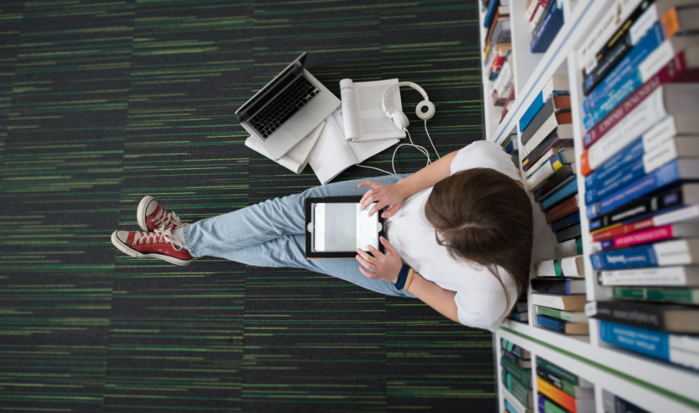 A shot taken from above a student sitting on a library floor while studying with an open laptop, a tablet and headphones