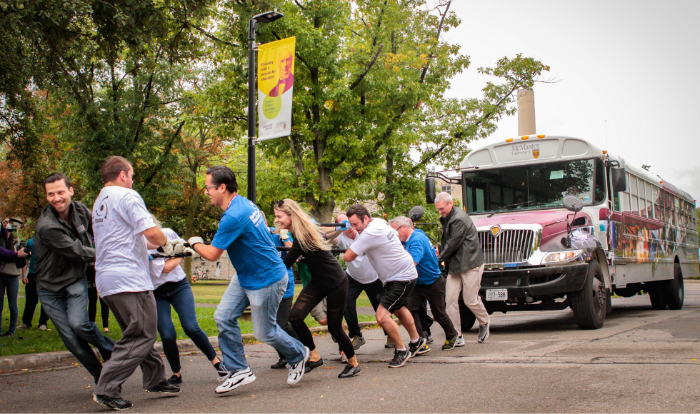 A group of people pulling a rope attached to a school bus that has McMaser University branding on it