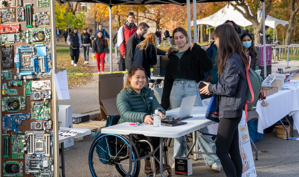 Students accept used tech at an outdoor booth. A student in a wheelchair smiles at a donor and beside her is a wall of circuitboards