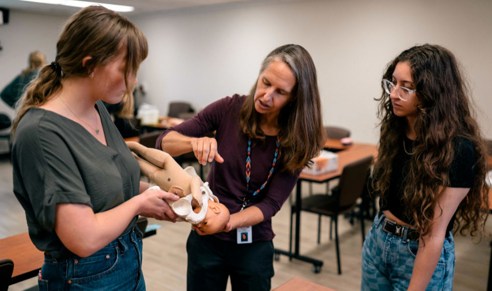 Three people looking down at an infant doll with its head inside a life-size model of a pelvic bone