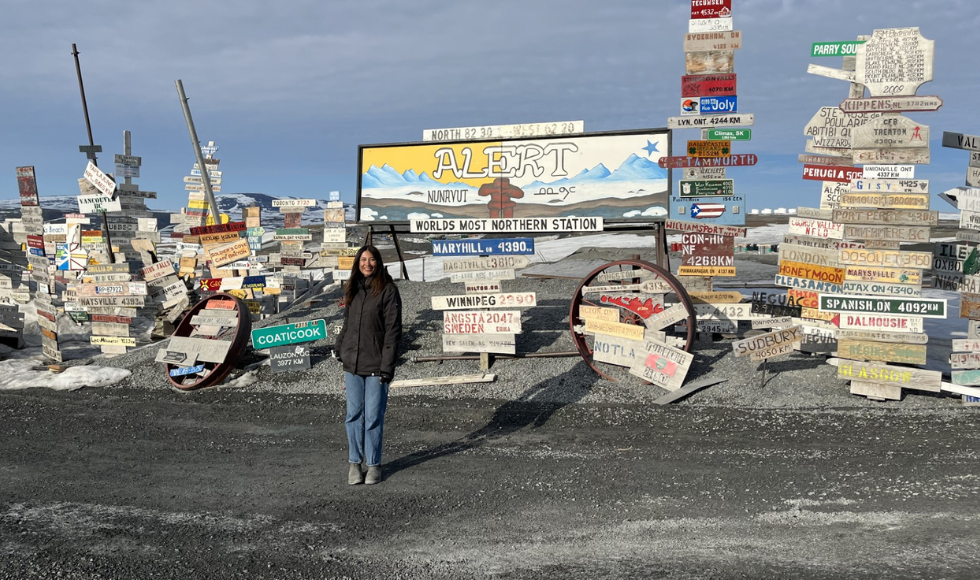 A woman standing in front of a sign that reads, ‘Alert Nunavut - world’s most northern station.’ Around the sign are dozens of other signs indicating how far Alert is from other cities around the world.