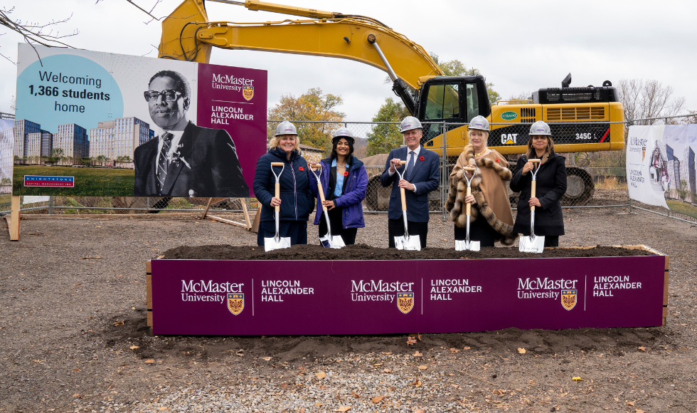 Five people with hard hats and shovels standing in front of a construction fence and a sign with a picture of Lincoln Alexander