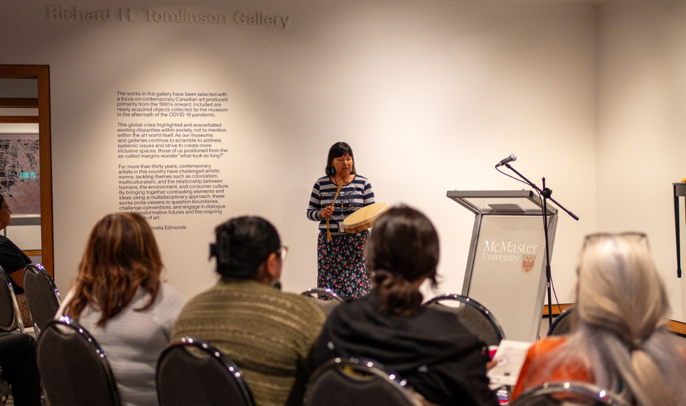 A woman beats a handheld drum with a stick in front of a seated audience in a room in the art gallery.