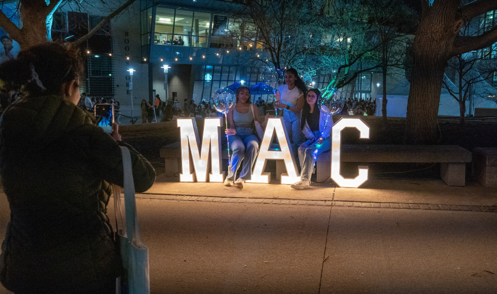 A person with their back to the camera taking a photo of three students posing with large lettering that spells out 'MAC
