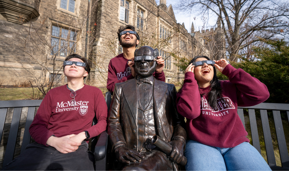 Three students in McMaster-branded clothing wearing total solar eclipse glasses. One of the students is holding a pair of solar eclipse glasses over the eyes of the statue of William McMaster.