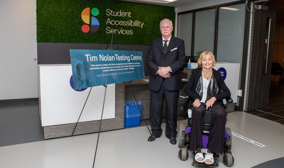 Tim Nolan is wearing a suit and standing next to his wife Kim, who is sitting in a power wheelchair. The couple is next to a sign that reads, “Tim Nolan Testing Centre,” along with a quote from Nolan – “Each person is unique, with their own passions and goals. McMaster is a place where individuality and leadership can be nurtured. Look for opportunities where you can make a positive difference in the lives of others.” In the background is the Student Accessibility Services logo.