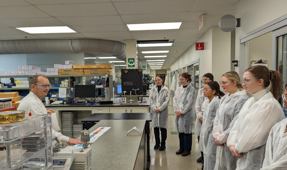 Image of students in lab coats, in a lab standing around listing to a man speak