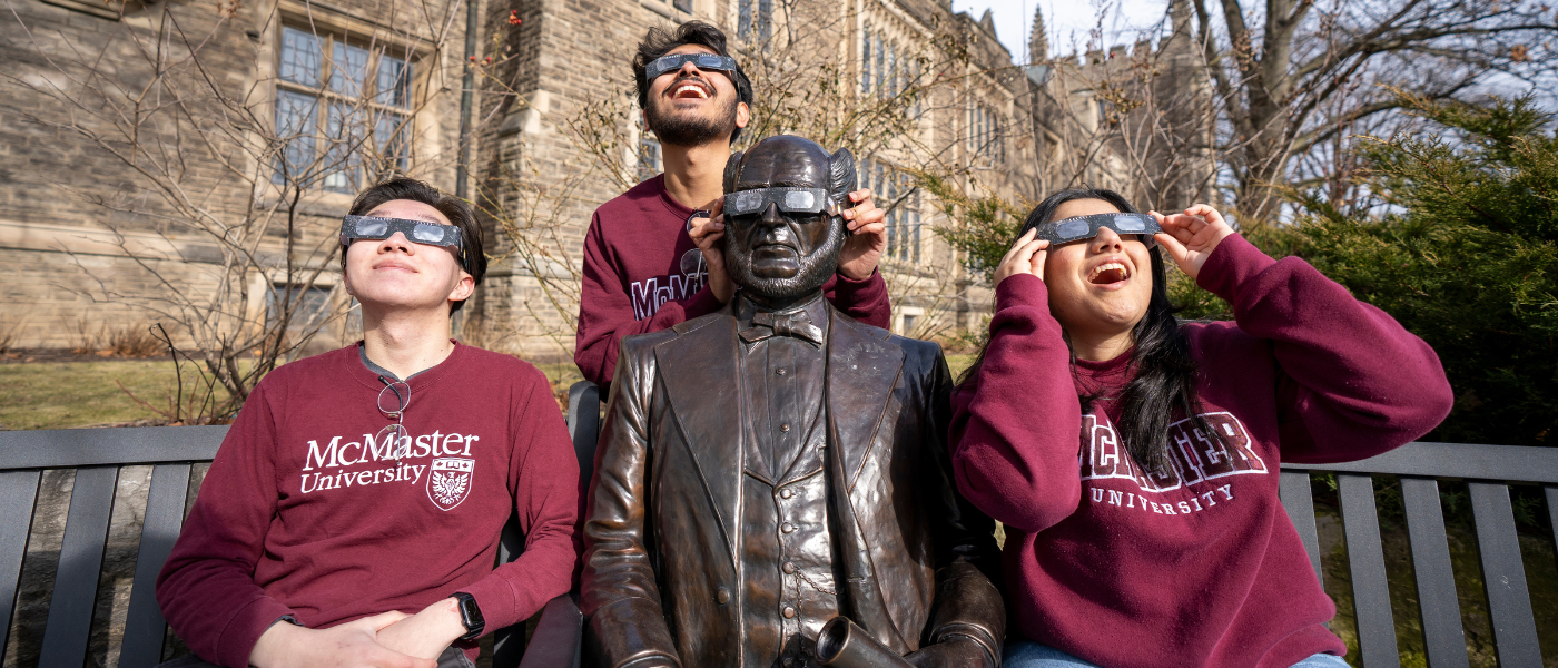 Three students in McMaster-branded clothing wearing total solar eclipse glasses. One of the students is holding a pair of solar eclipse glasses over the eyes of the statue of William McMaster.