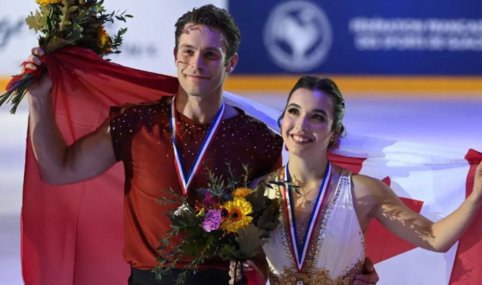 Image of Trennt Michaud standing beside his partner Lia Pereira they both have medals around their necks, and flowers in their hand, while holding the Canadian flag behind their backs.