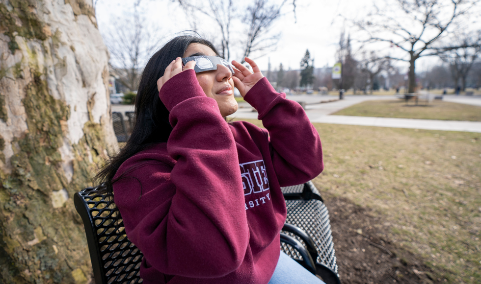 A student in a McMaster sweatshirt wearing solar eclipse glasses and looking up at the sky