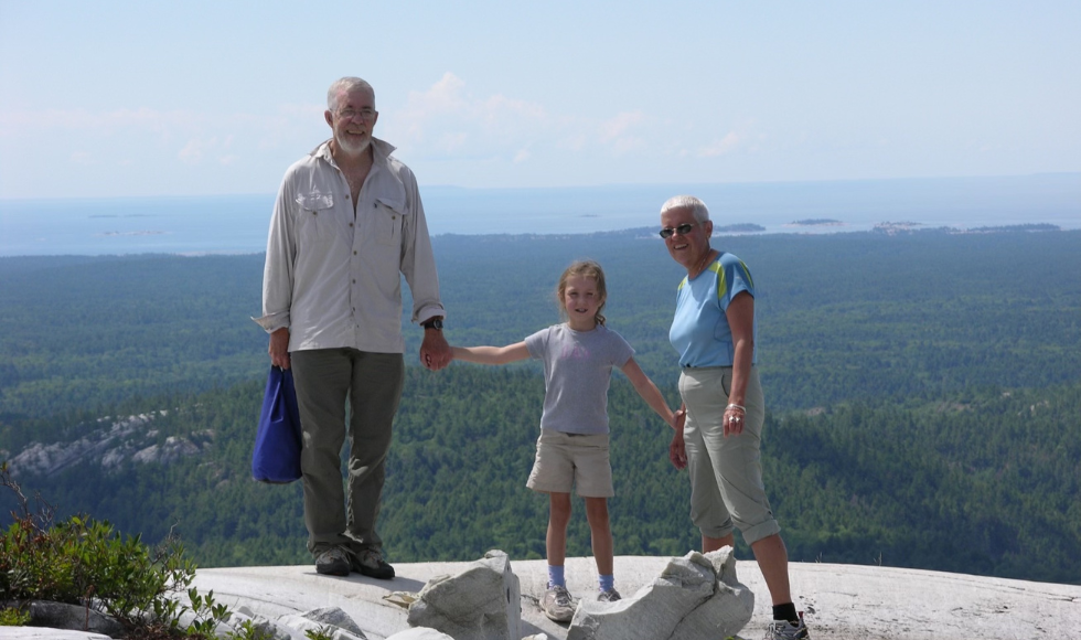 An older image of a young Emma and her grandparents holding her hands as they stand at the top of the mountain and the park