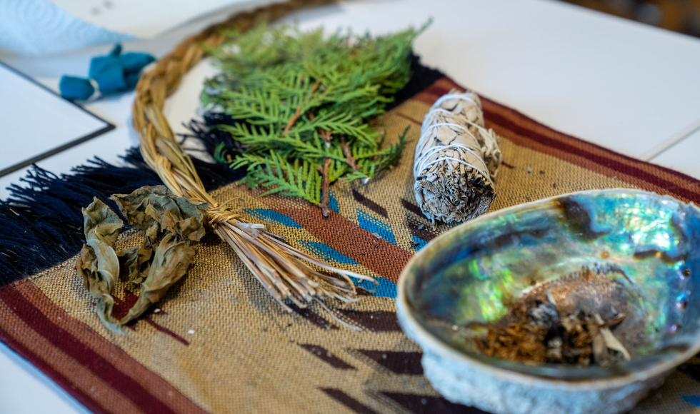 Materials that would be used in a smudging ceremony, including a smudge bowl and some cedar leaves, lie on a table