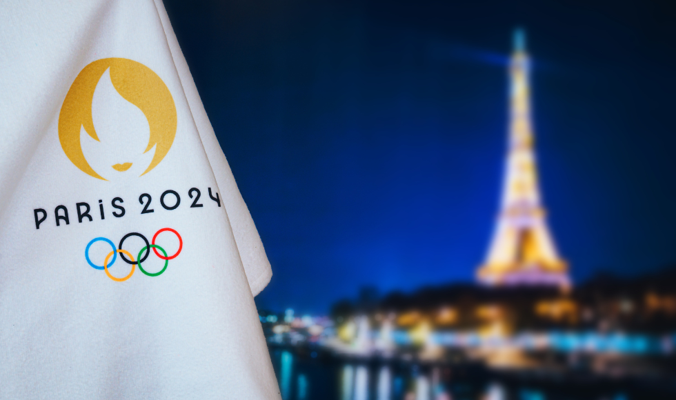 A portion of of a white flag with the Olympic logo on it and text that reads, 'Paris 2024.' The Eiffel Tower out of focus can be seen in the distance.