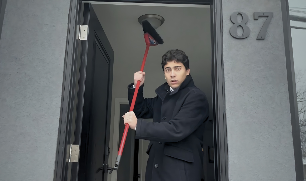 A man with a scared look on his face holding up a red broom while standing in the doorway of a home