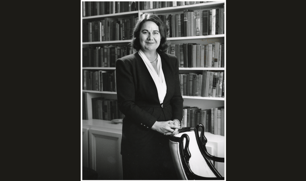 Black and white portrait of Geraldine Kenney-Wallace standing in front of a book-lined wall, her hands resting on a chair.