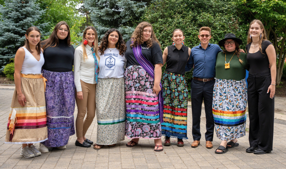 Nine people standing outdoors arm-in-arm smiling at the camera