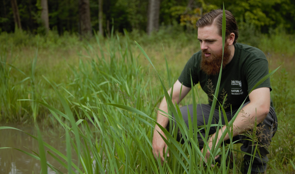 Noah Stegman crouched down in tall grasses looking at a body of water.