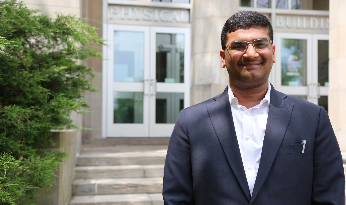 Smiling Udbhav Ram wearing a blazer and a white shirt in the foreground, with Burke Science Building behind him.