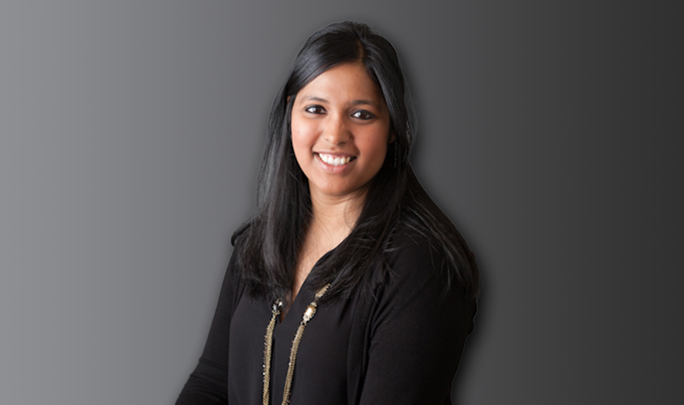 A woman in a black long sleeve shirt with a gold necklace and dark hair sits in front of a grey background.