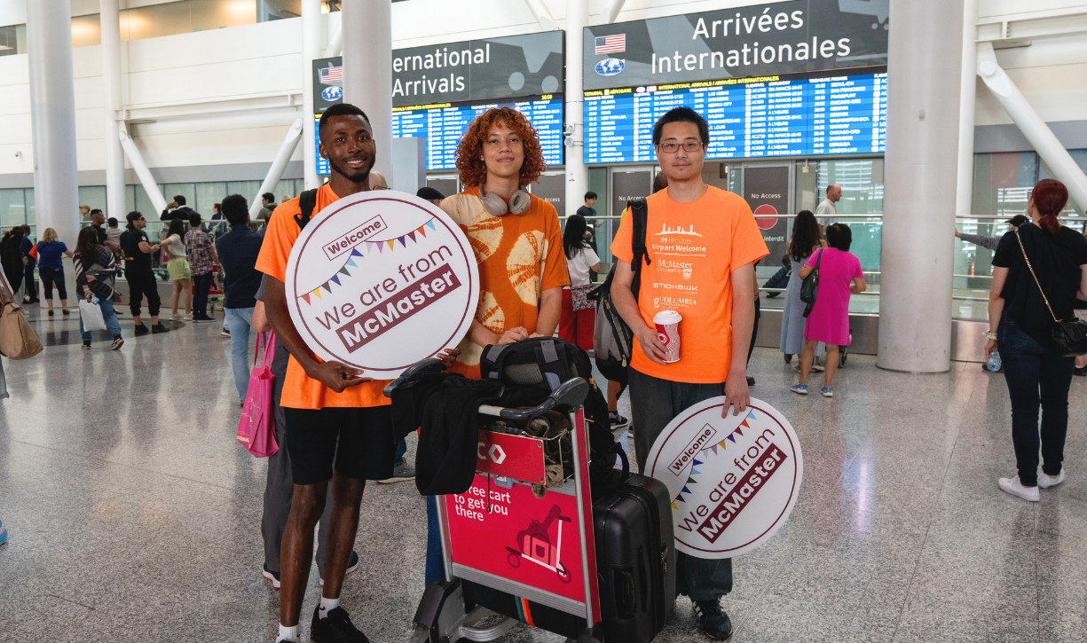 An arriving international student stands with two Airport Welcome volunteers holding We are From McMaster signs inside Pearson Airport