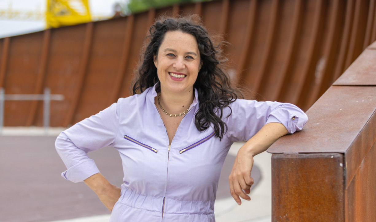 A smiling woman with long dark hair in a lilac shirt, seen from waist up, outdoors.