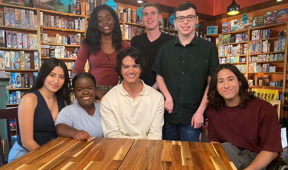 Group of 7 young adults, some seated at a table, some standing, against the backdrop of a board games cafe.