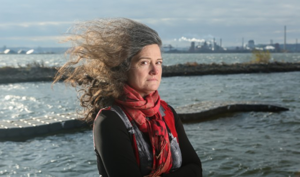 Kelly Wolf looking sideways at the camera, with the wind blowing her hair straight up behind her, against a backdrop of waves in the sea or a lake.