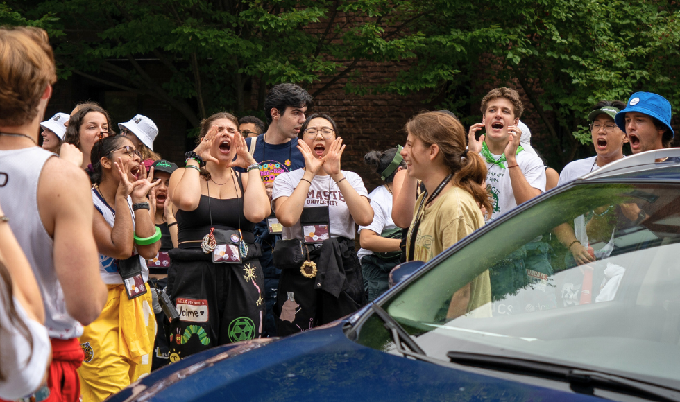 A crowd of students cheers while a student standing beside a car smiles back at them