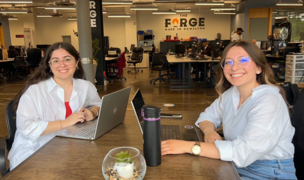 Two students sitting at a desk smiling at the camera. The logo of The Forge, McMaster's business incubator can be seen on the wall behind them.