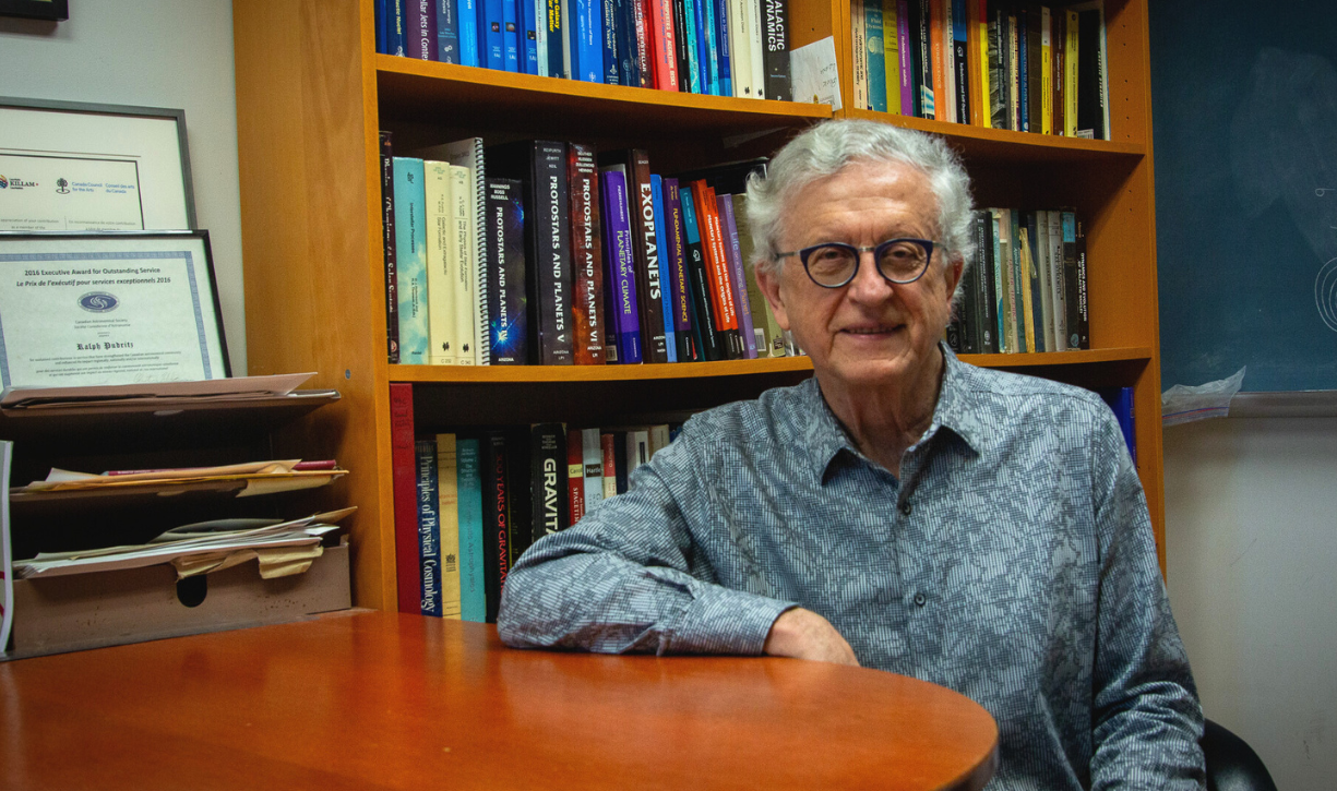 Ralph Pudritz in a blue dress shirt sits in an office with a bookshelf, certificates, and blackboard with equations in the background.