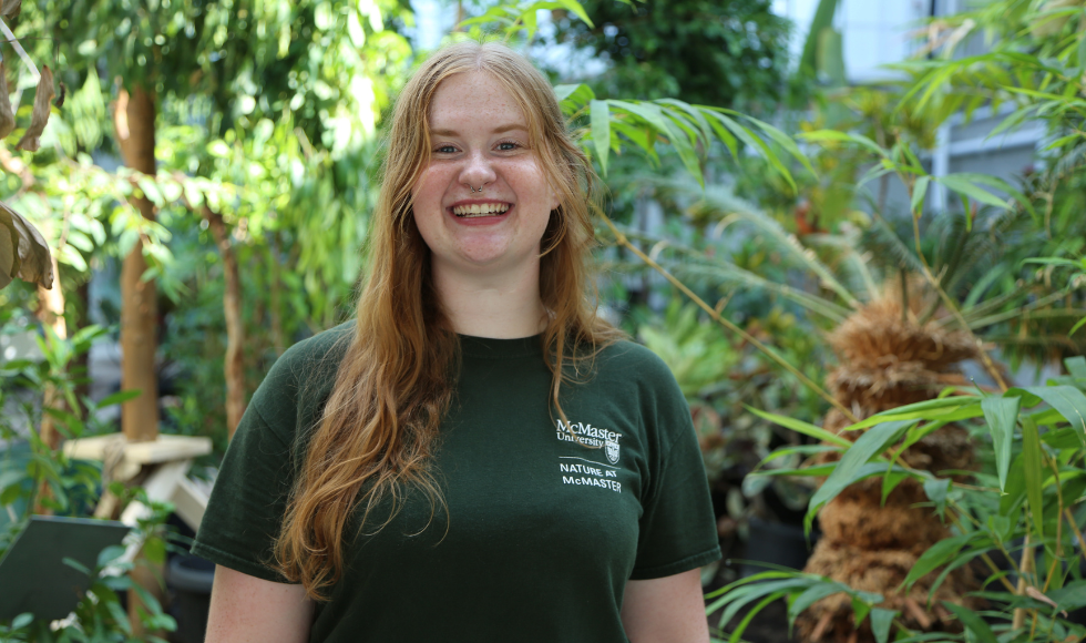 Renee Twyford, wearing a Nature at McMaster T shirt, smiles widely in the new greenhouse against a backdrop of tropical greenery.