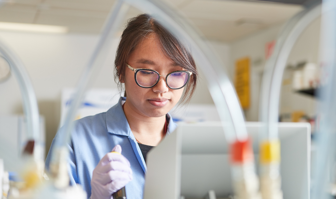 A person wearing a lab coat and gloves in a lab, holding lab equipment, looking down.