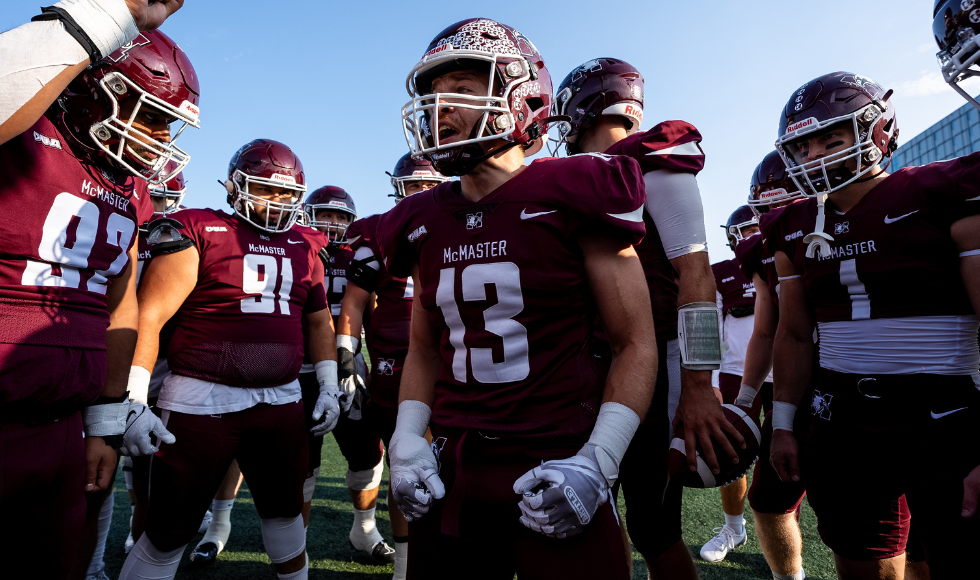 McMaster Marauders at their home opener game. A group of a dozen football players wearing maroon helmets and jerseys gather in a huddle.