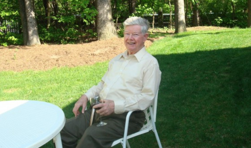 Alan Hitchen sitting outdoors on a patio chair smiling at the camera. There is a round white table in front of him and he is holding a glass of red wine in one hand.