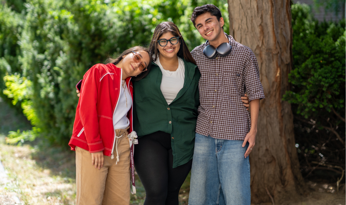 Three smiling students standing with their arms around each other's shoulders or waists in front of greenery and tree on campus. The one on the left is leaning her head on the centre student.