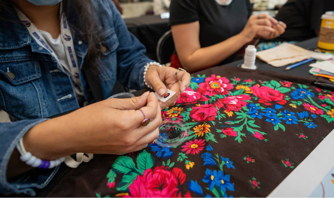 A closeup of a person's hands as they do very precise and intricate beading at a table with a colourful tablecloth on it.