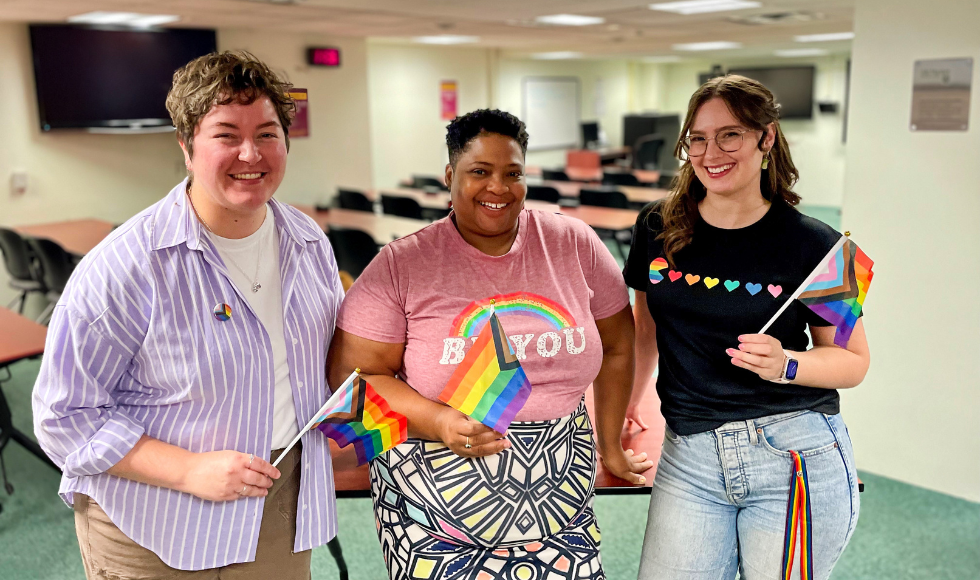 Three people standing and smiling at the camera. They are each holding a 2SLGBTQ+ flag.