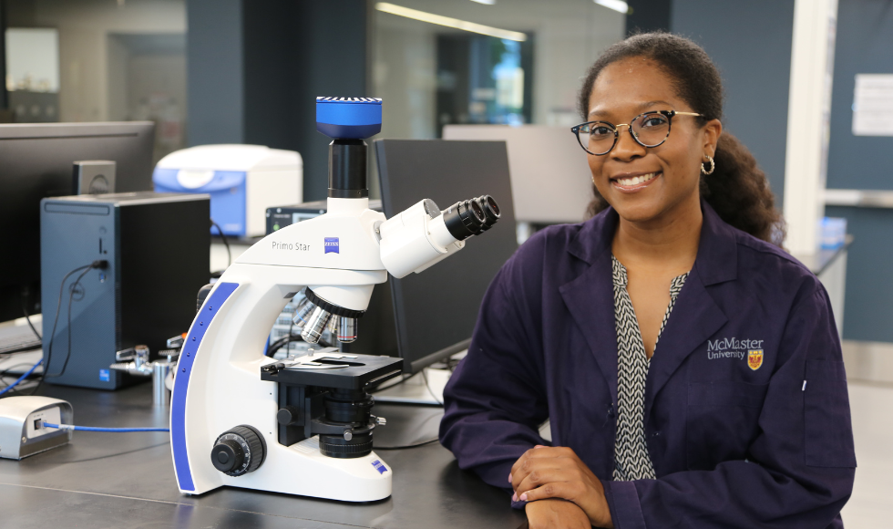 Shaiya Robinson seated beside a microscope smiling directly at the camera. She is wearing a blue lab coat that has the McMaster University logo on it.