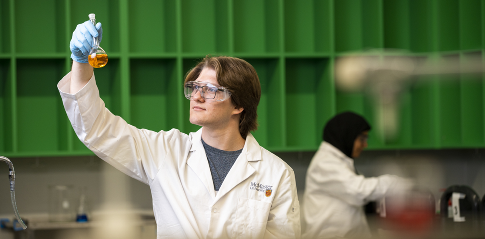 A student standing in a laboratory wearing personal protective gear and holding up a vial that has an amber liquid in it.