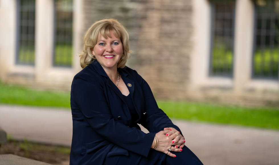 A woman in a navy suit with blond hair, faces camera with a smile. She is sitting on a bench with stonework and green grass behind her.