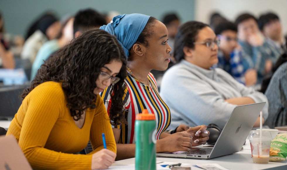 Three students sit at a desk in the foreground. Behind them, out of focus, are more students seated at desks listening to someone out of view of the camera.