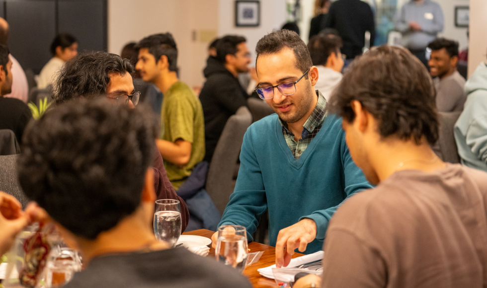 A group of students sitting at a dining table. In the background other groups of people around dinign tables can be seen.