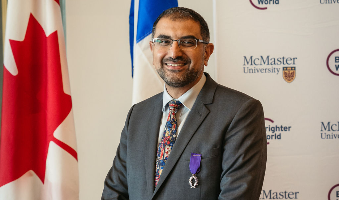 Hatem Zurob, smiling in a suit and with an Order of the Academic Palms medal pinned to his chest, against a backdrop of the McMaster University banner and the Canadian flag.